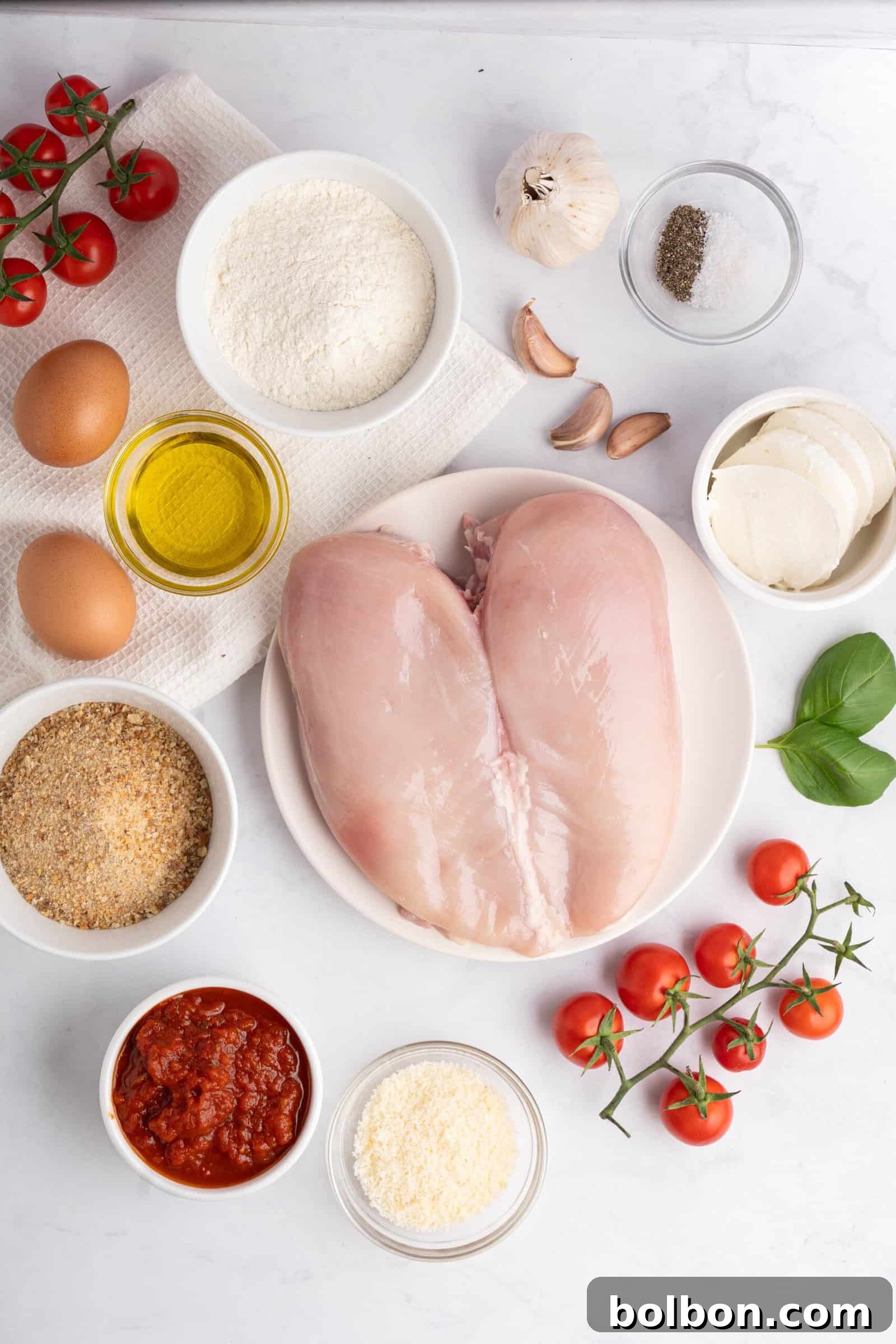 Homestyle Baked Chicken Parmigiana 3 A flatlay of ingredients for a meal: raw chicken breasts, eggs, flour, breadcrumbs, olive oil, marinara sauce, mozzarella, grated cheese, cherry tomatoes, garlic cloves, black pepper, and fresh basil on a white surface.