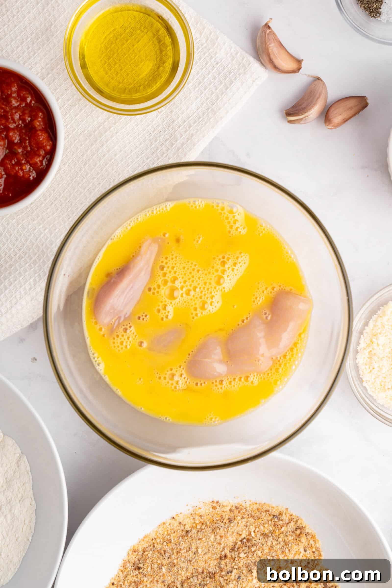 Homestyle Baked Chicken Parmigiana 4 Raw chicken strips are dipped in a bowl of beaten eggs. Surrounding the bowl are ingredients including olive oil, garlic cloves, tomato sauce, flour, breadcrumbs, and seasonings on a white surface.