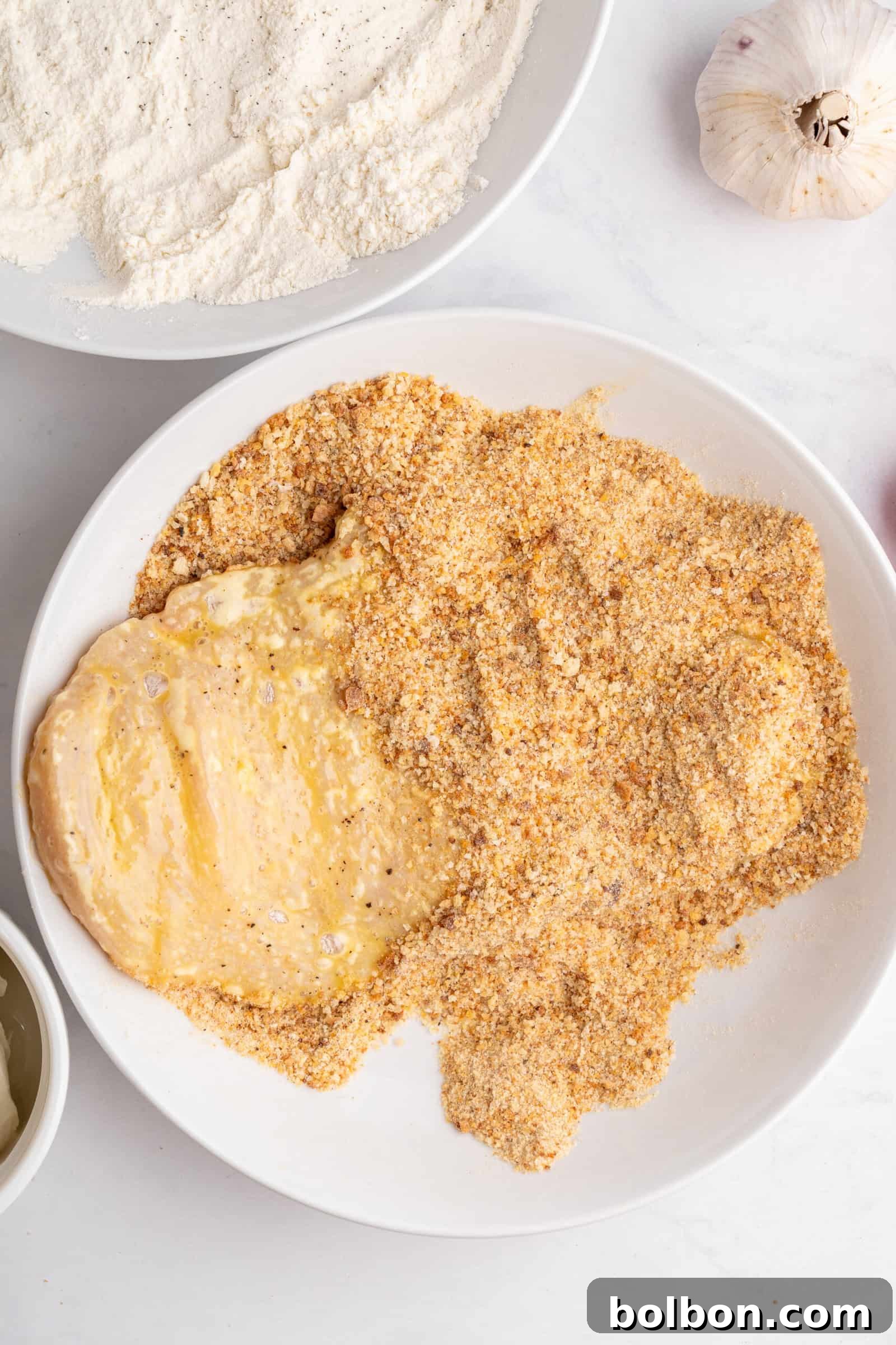 Homestyle Baked Chicken Parmigiana 5 A bowl of chicken breast being coated in seasoned breadcrumbs, with flour and garlic nearby. The chicken appears partially covered, ready for cooking.
