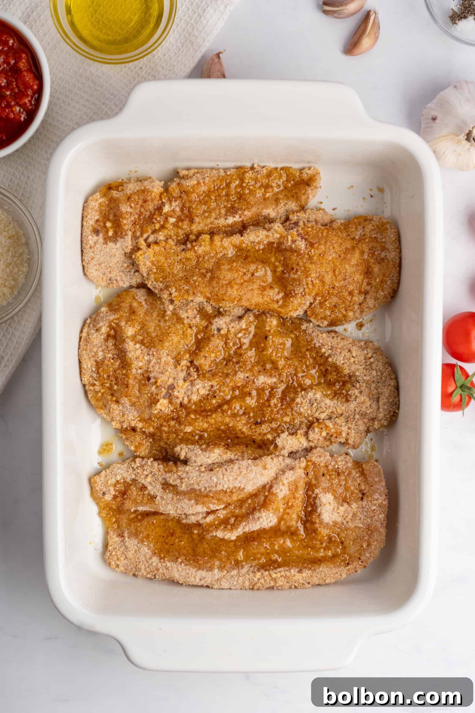 Homestyle Baked Chicken Parmigiana 6 Raw breaded chicken fillets are placed in a white baking dish on a marble surface, ready for cooking. Surrounding the dish are ingredients like olive oil, tomato sauce, garlic, and parmesan cheese on a white towel.