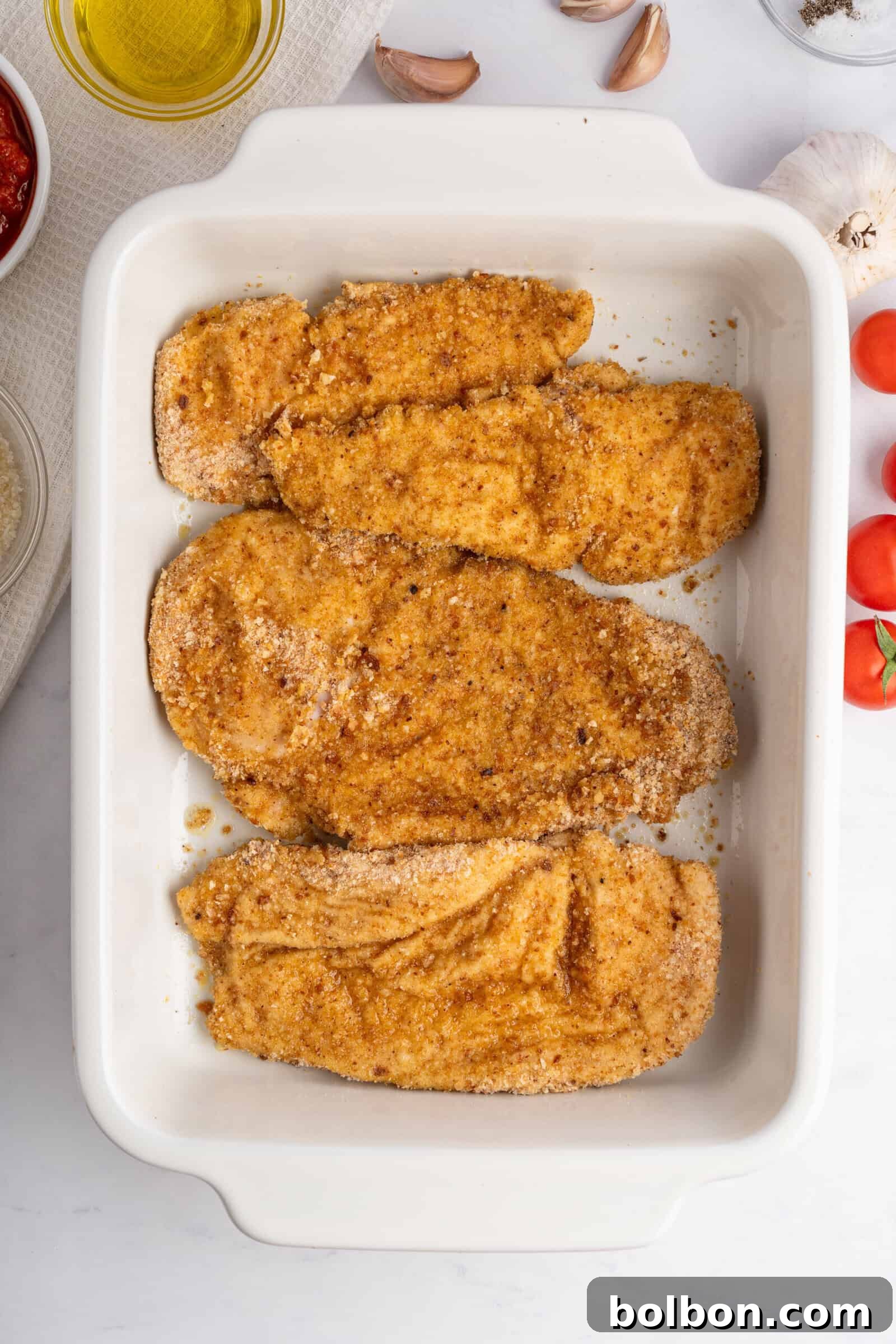 Homestyle Baked Chicken Parmigiana 7 Top view of a white baking dish containing four breaded chicken cutlets. The cutlets are seasoned with spices and breadcrumbs. Ingredients like cherry tomatoes, garlic, and olive oil are nearby on a marble countertop.