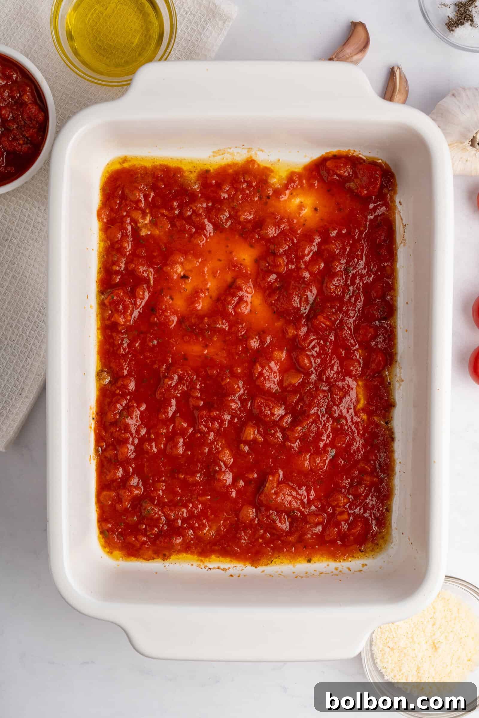 Homestyle Baked Chicken Parmigiana 8 A white rectangular baking dish filled with tomato sauce on a light surface. Nearby are ingredients including olive oil, garlic cloves, and grated cheese, suggesting preparation for a dish.