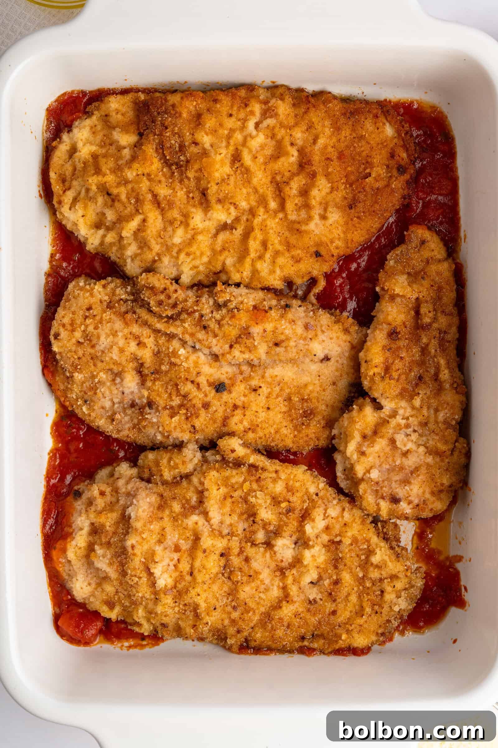 Homestyle Baked Chicken Parmigiana 9 A white baking dish containing four breaded and browned chicken cutlets on a bed of red tomato sauce.