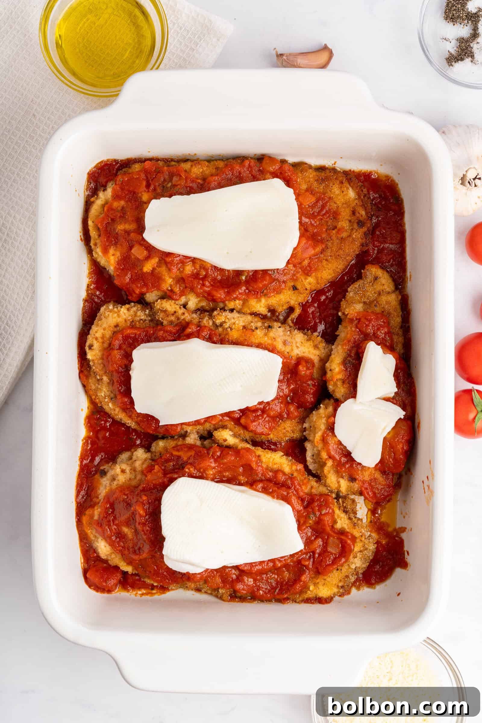 Homestyle Baked Chicken Parmigiana 10 A baking dish containing breaded chicken breasts topped with tomato sauce and slices of cheese. Surrounding the dish are ingredients like tomatoes, garlic, mushrooms, a bowl of grated cheese, and a small dish of olive oil.