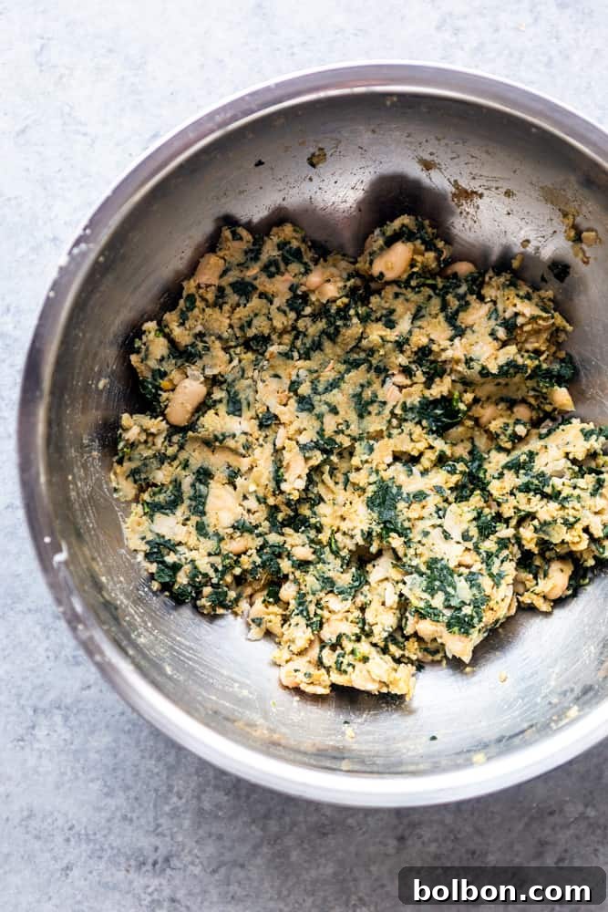 A close-up view of the perfectly mixed white bean and kale patty batter in a gleaming silver bowl, showcasing its consistent texture and vibrant green flecks of kale.