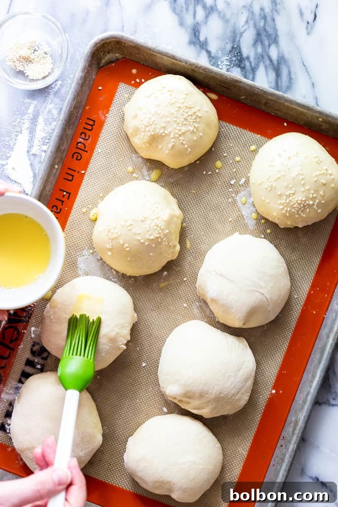 Hands brushing an egg wash mixture over unbaked hamburger buns, topped with sesame seeds, ensuring a beautiful golden finish during baking.