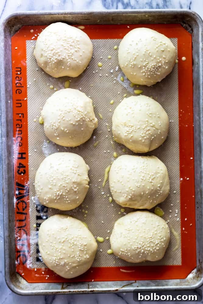Eight perfectly shaped unbaked hamburger buns arranged on a baking sheet, topped with sesame seeds, glowing golden from the egg wash, waiting to be baked.