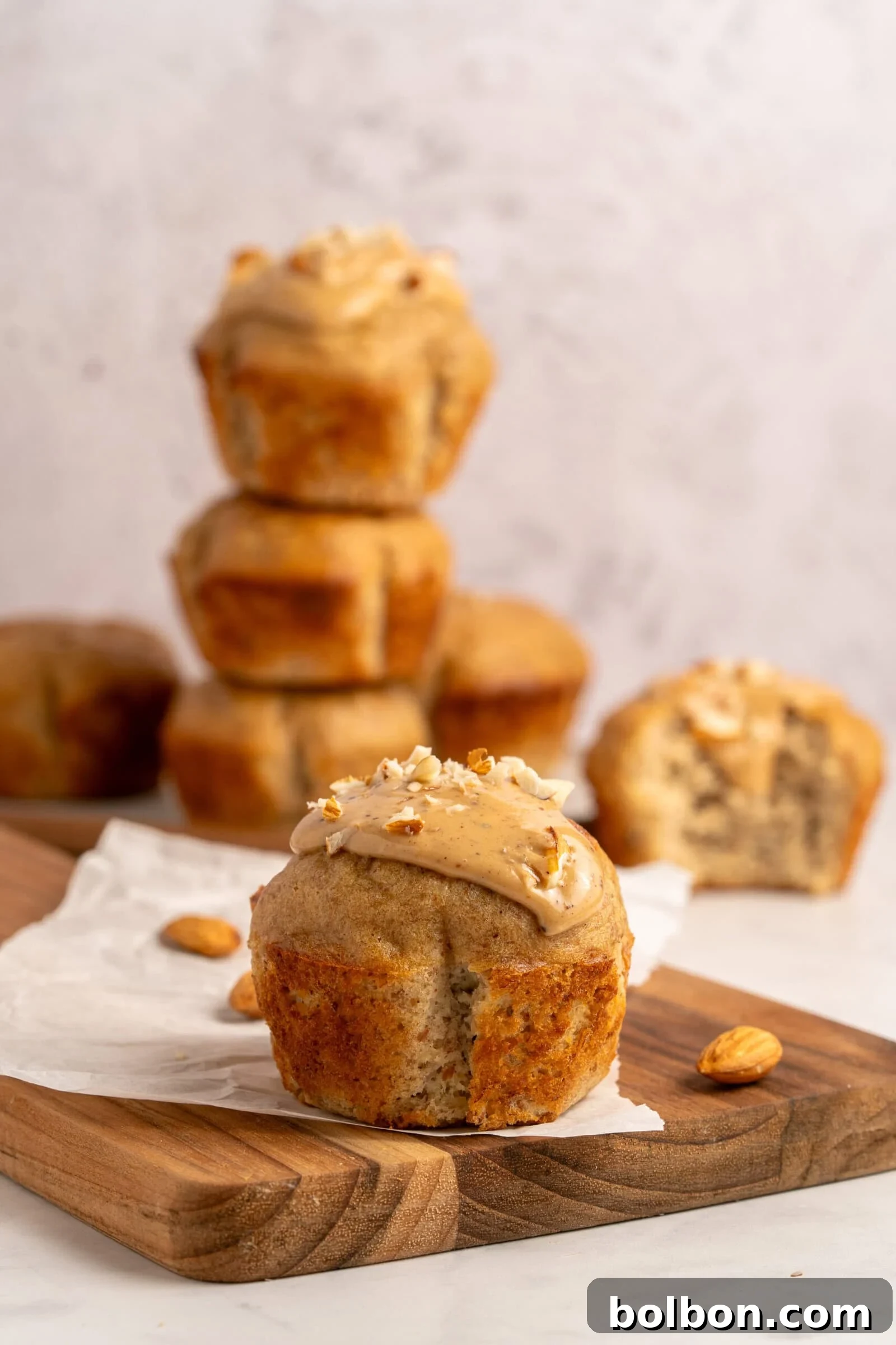 Wholesome Almond Muffins 2 A close-up of a muffin topped with frosting and chopped nuts on a wooden board. In the background, more muffins are stacked, with almonds scattered around. The scene has a light, neutral background.