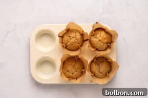 A four-cup muffin tin holding four baked muffins in brown parchment paper liners, with two empty cups, on a light marble surface.
