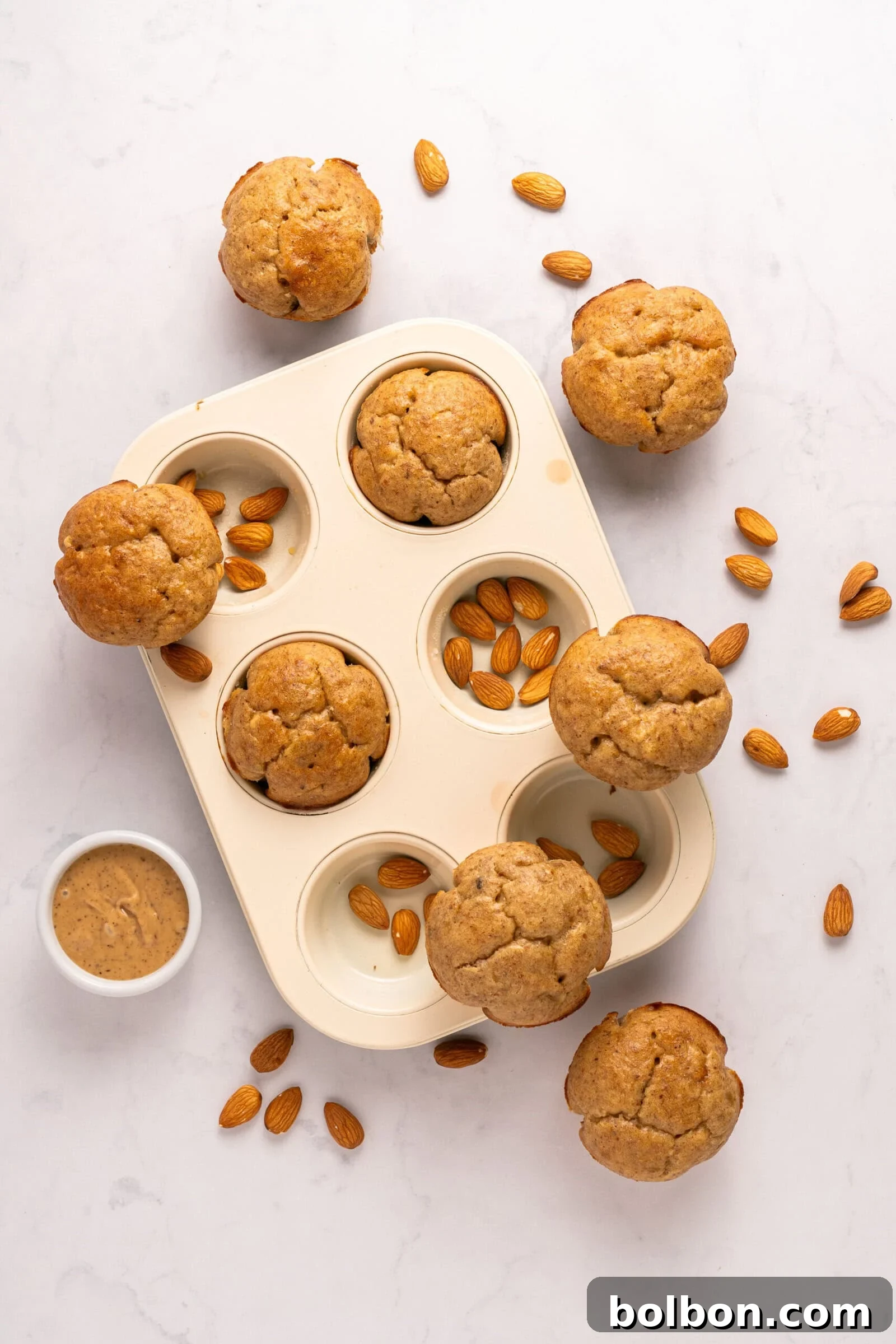 Wholesome Almond Muffins 7 A white muffin tray with six almond muffins, surrounded by scattered whole almonds and a small bowl of almond butter on a white marble surface.