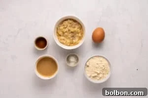 Top-down view of six baking ingredients on a white surface: mashed banana, an egg, flour, baking powder, peanut butter, and vanilla extract, each in separate bowls or containers.