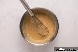 A metal whisk rests inside a stainless steel bowl filled with a speckled, creamy batter on a light-colored marble surface.