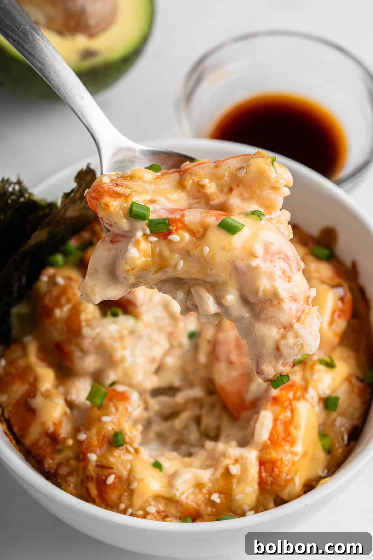 A spoon holds a creamy shrimp over a white bowl filled with similar shrimp, garnished with chopped green onions and sesame seeds. A small dish of soy sauce and an avocado are in the background.