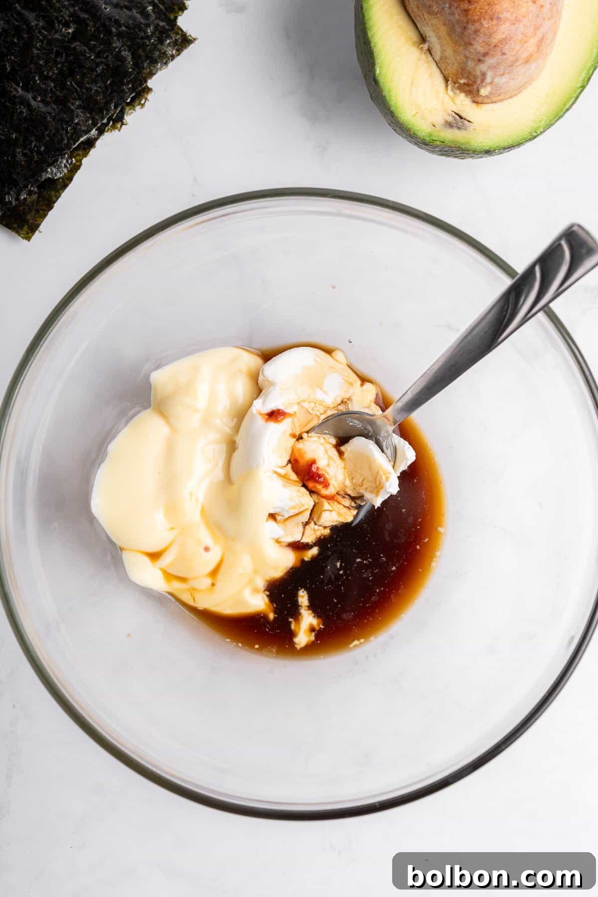 A glass bowl containing mayonnaise, soy sauce, and a small amount of mustard, with a spoon inside. Nearby are a halved avocado and a piece of seaweed on a light-colored surface.