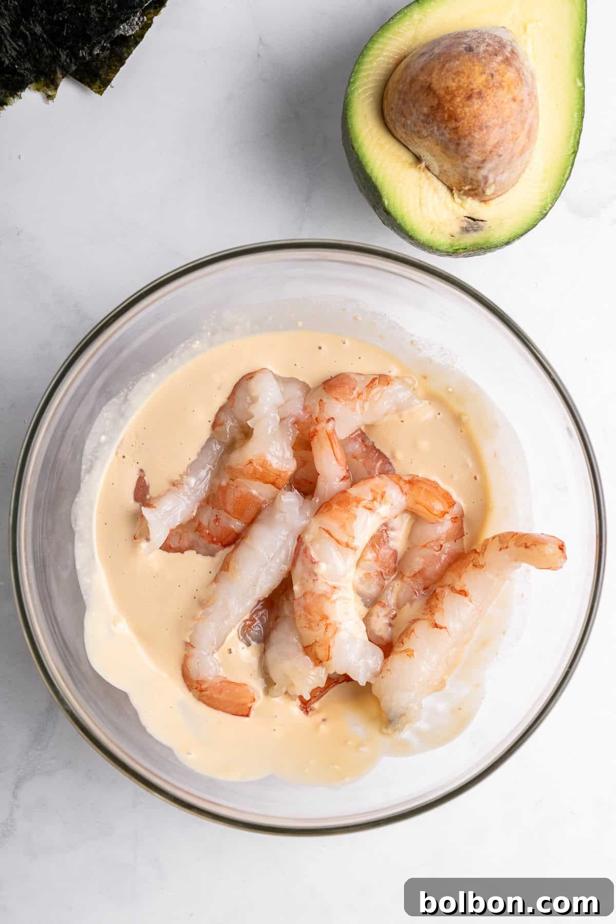 A glass bowl filled with shrimp partially submerged in a creamy sauce. Next to the bowl is a half-avocado with the seed intact. A piece of seaweed is also visible in the top left corner of the image.