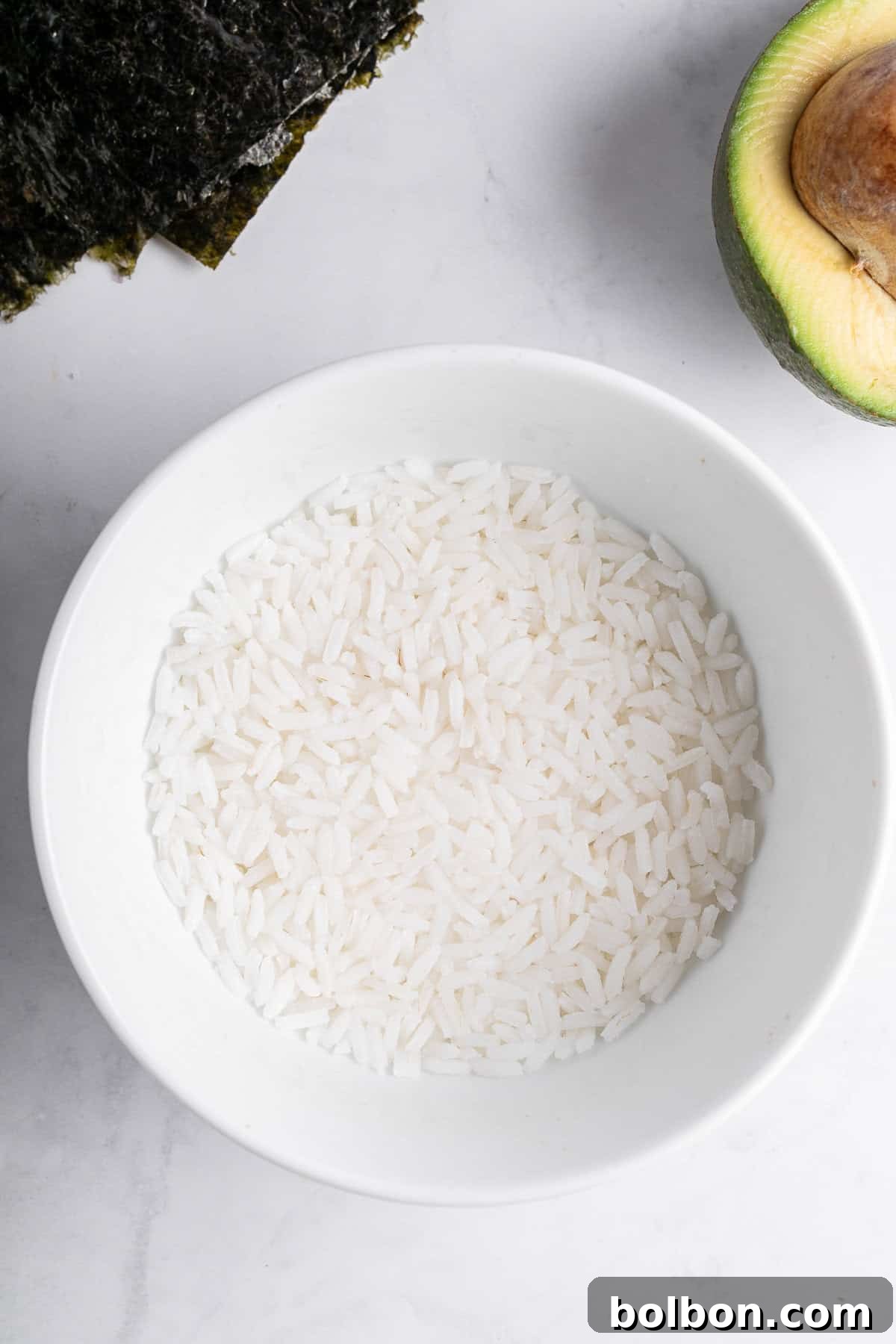 A white bowl filled with uncooked white rice is placed on a marble surface. To the side, there is a halved avocado with the pit visible and a sheet of nori seaweed.