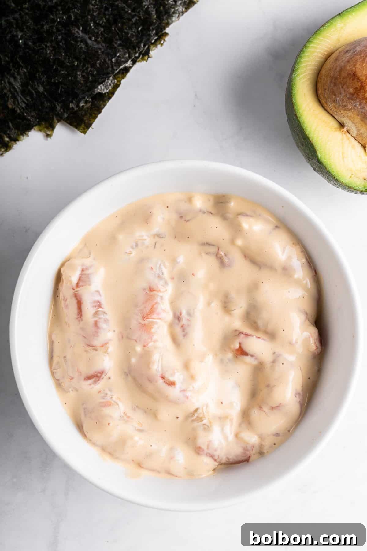 A white bowl filled with a creamy mixture containing chunks of pink seafood, possibly shrimp or crab, is placed on a white marble surface. A partial view of an avocado and sheets of seaweed are visible nearby.