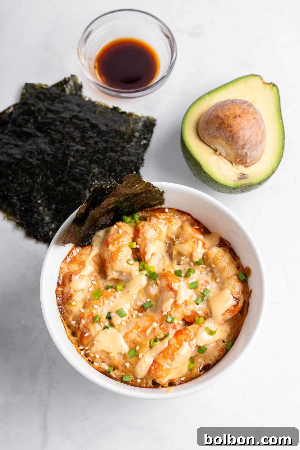 A bowl of creamy baked sushi with shrimp, topped with green onions and sesame seeds. Nori sheets, soy sauce in a small bowl, and a halved avocado are placed nearby on a light surface.