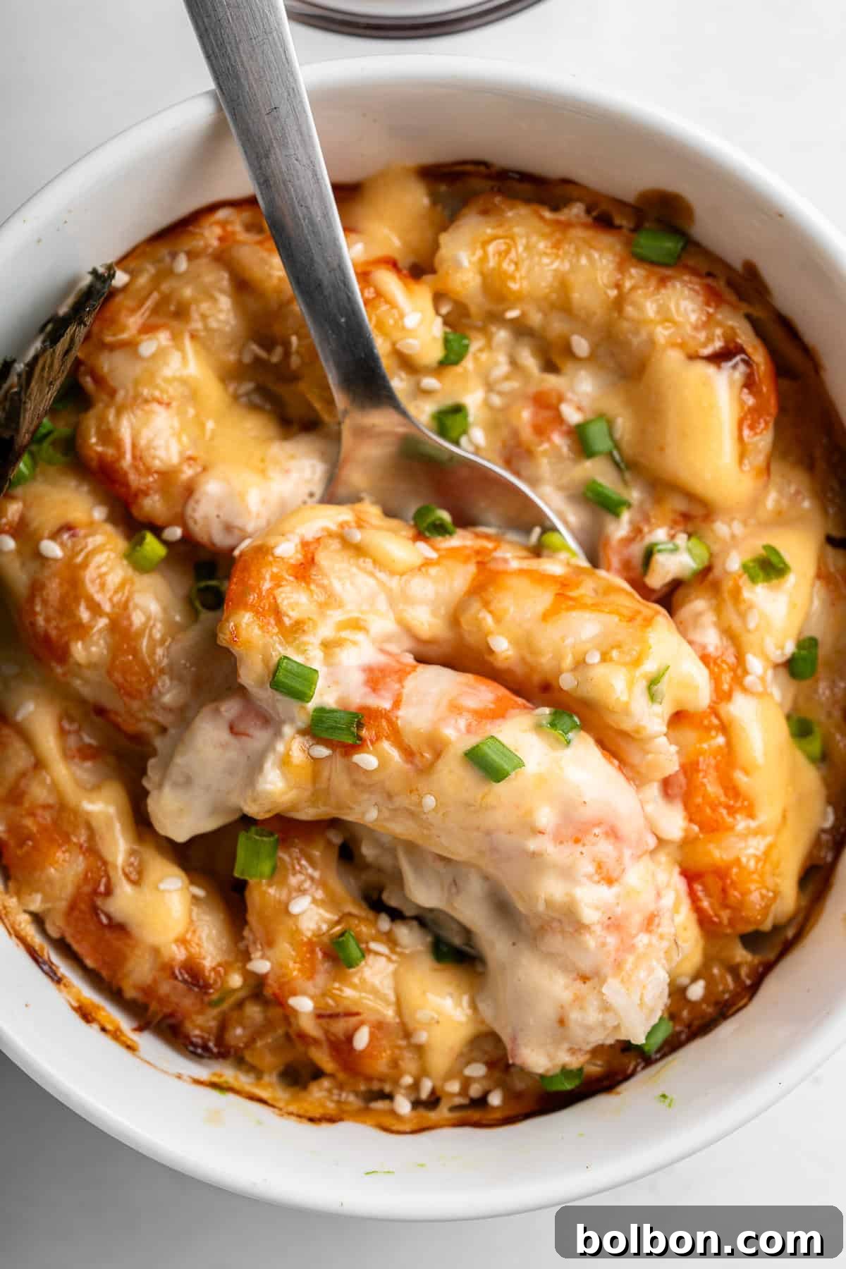 A close-up of a creamy Sushi bake recipe dish in a white bowl. The shrimp are coated in a cheese sauce, garnished with chopped green onions and sesame seeds. A spoon is partially submerged in the dish, showing the rich texture.