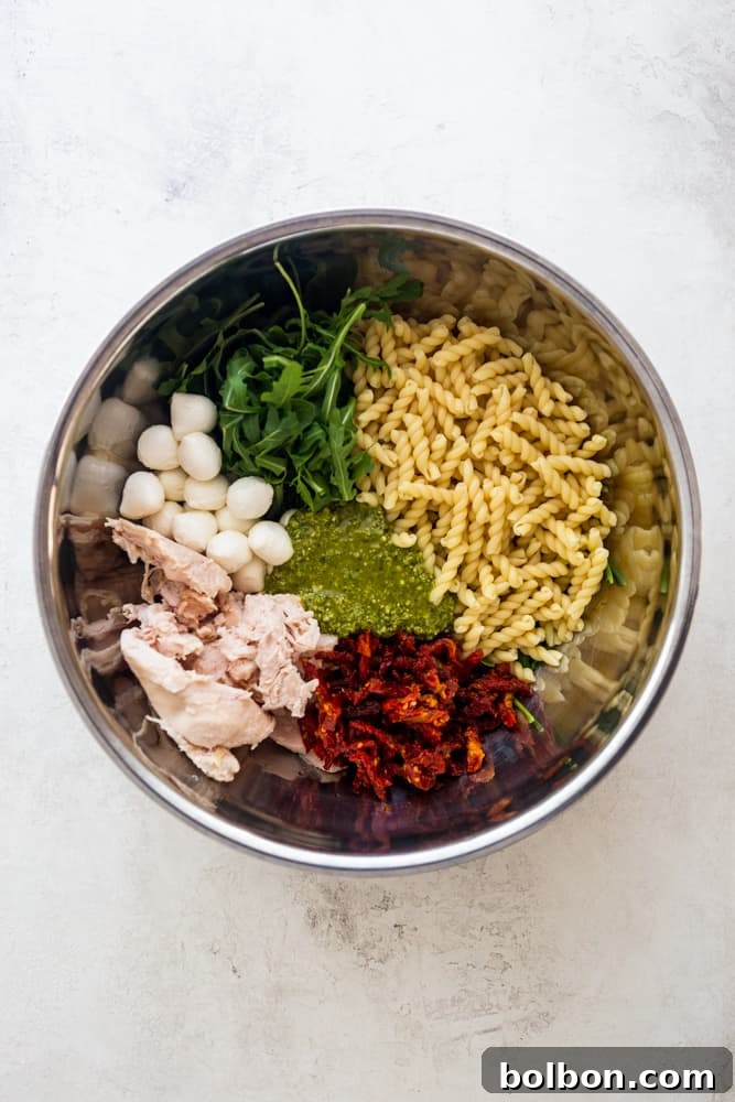 Fresh ingredients for chicken pesto pasta salad including cooked pasta, vibrant arugula, small mozzarella balls, shredded cooked chicken, sun-dried tomatoes, and a jar of pesto, all in a large stainless steel bowl