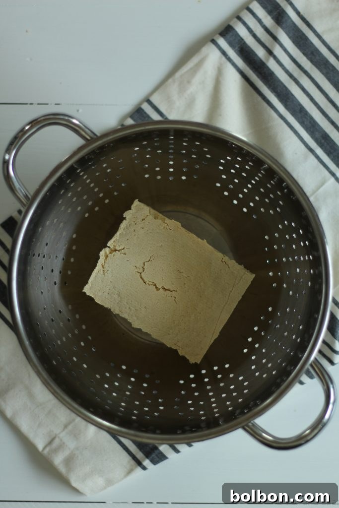 Perfect Tofu Secrets 5 DIY tofu press setup with a block of tofu in a colander, topped with paper towels and heavy bowls, draining water.
