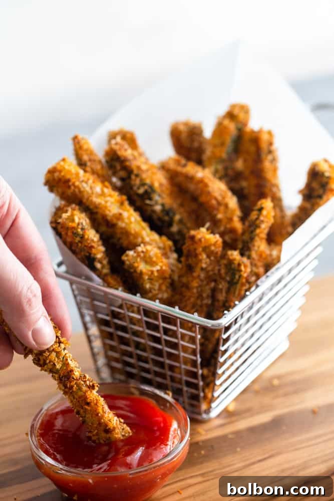 Golden Baked Zucchini Sticks 8 A basket of crispy baked zucchini fries, with one fry being dipped into a small bowl of ketchup.