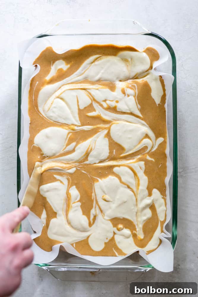 A knife gently swirling cream cheese into pumpkin batter in a baking dish, creating a marbled pattern for the bars. Keywords: swirling technique, marbling cream cheese, pumpkin dessert preparation, visual appeal.