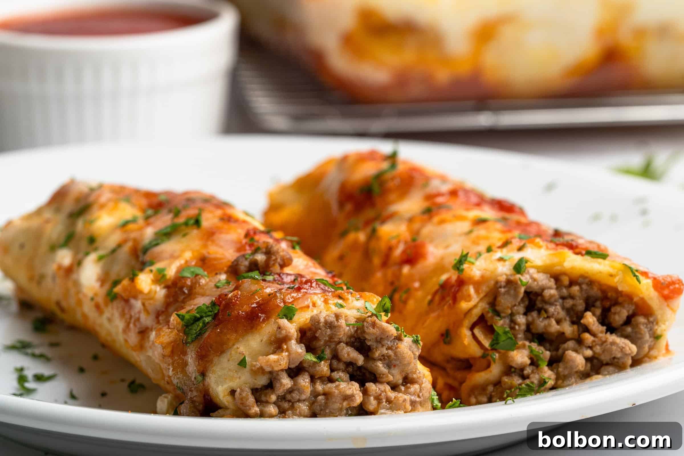 Crepe Lasagna Bake 2 Two beef-stuffed cannelloni topped with melted cheese and herbs on a white plate. A dish of sauce is in the background.
