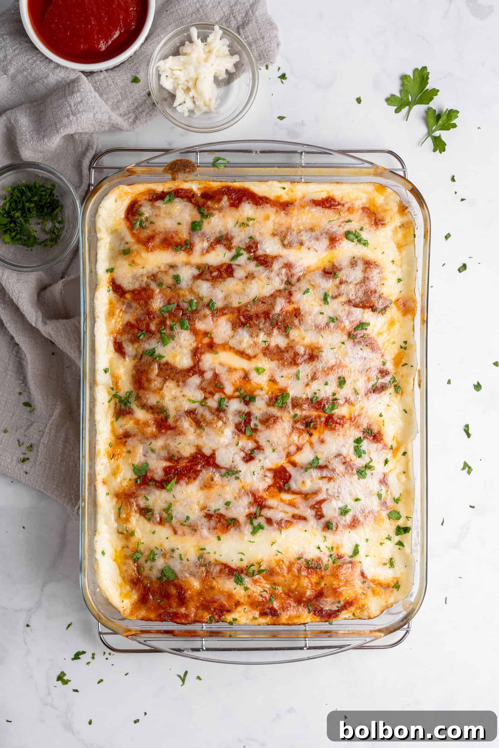 Crepe Lasagna Bake 11 A glass baking dish filled with freshly baked lasagna topped with melted cheese and sprinkled with chopped parsley. Ingredients like grated cheese and tomato sauce are nearby on a white marble surface.