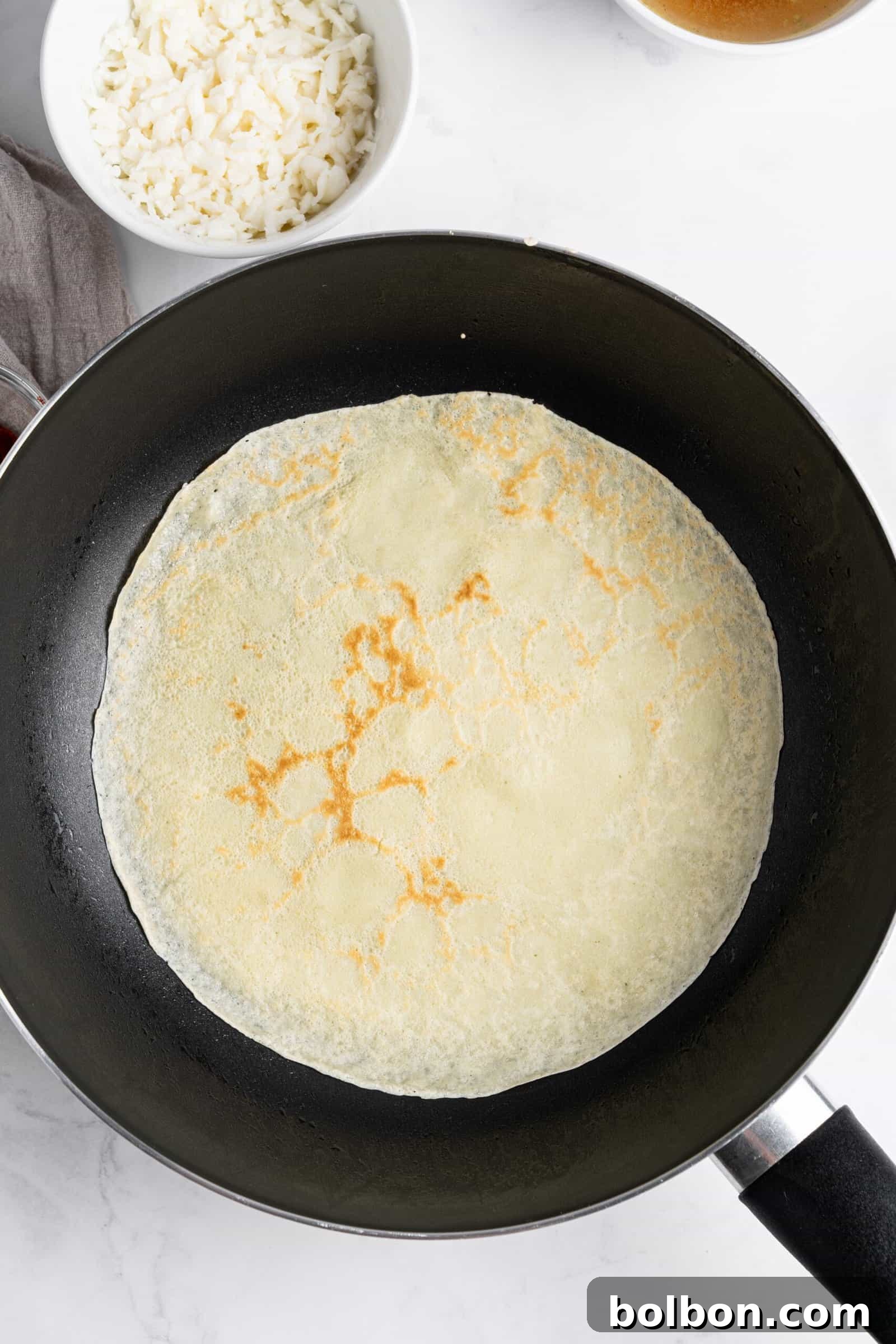 Crepe Lasagna Bake 4 A crepe cooking in a black non-stick pan on a white countertop. Nearby, a bowl of white rice is partially visible.