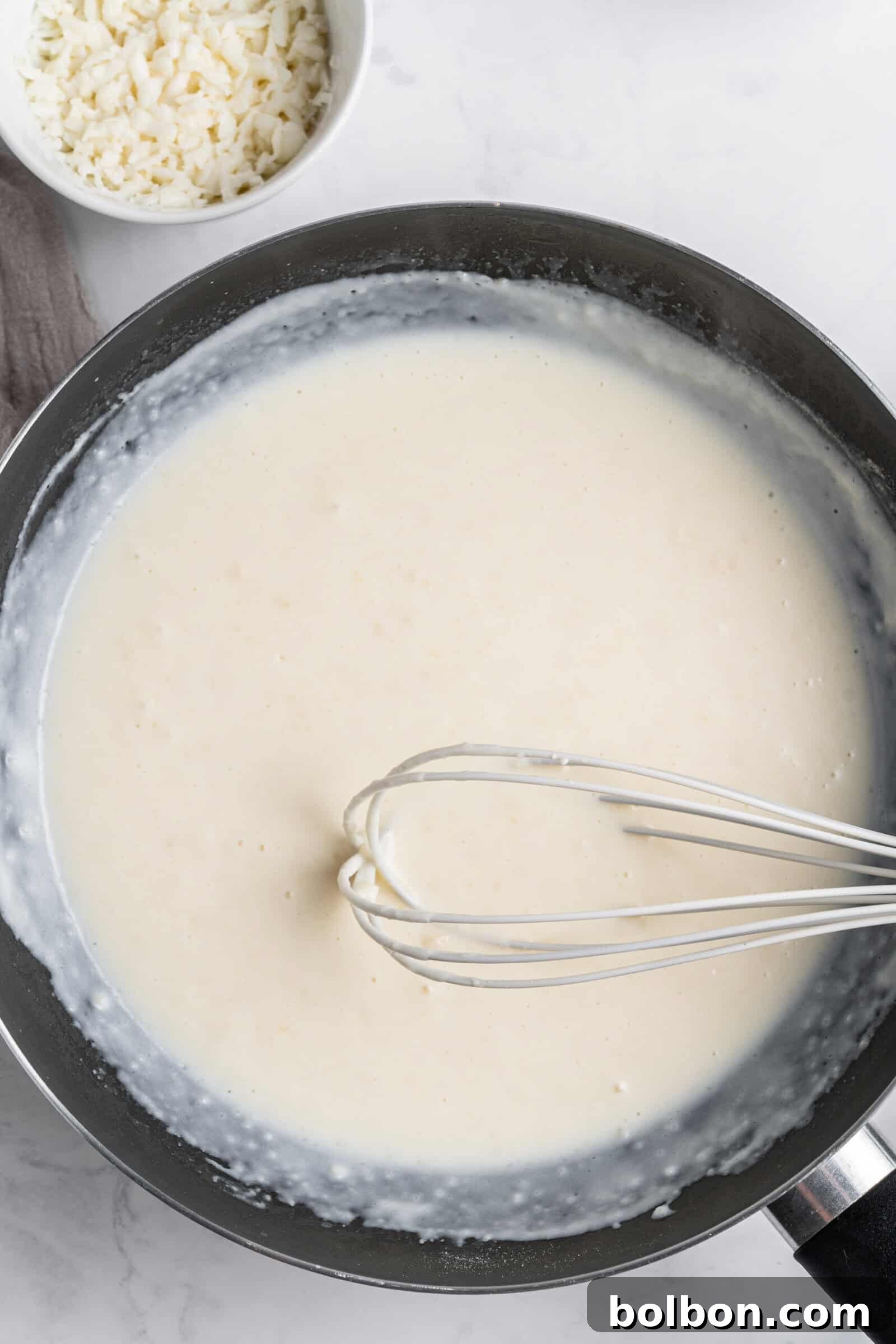 Crepe Lasagna Bake 5 A whisk stirring a creamy white sauce in a black pan on a marble surface. A small white bowl containing grated cheese is placed nearby.