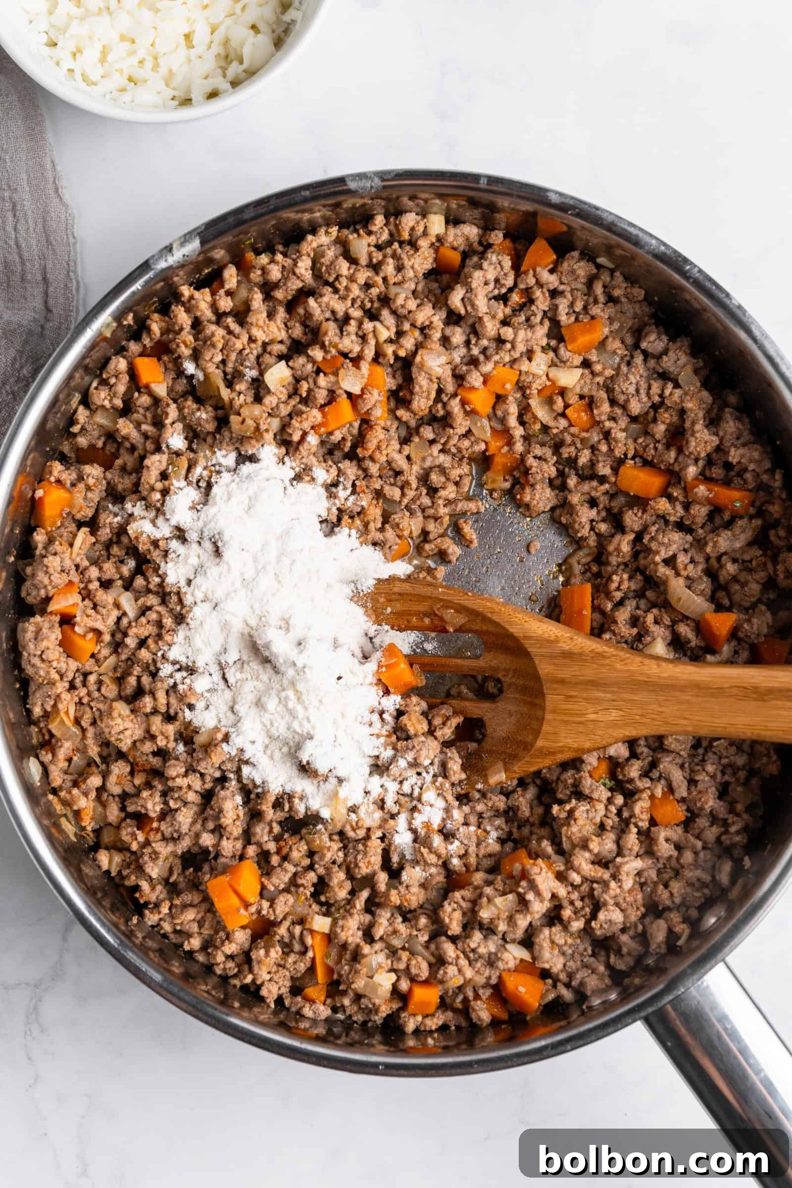 Crepe Lasagna Bake 7 A skillet filled with cooked ground meat and chopped orange bell peppers, topped with a pile of flour. A wooden spatula rests inside the skillet, and a small bowl of white shredded cheese is visible in the background.