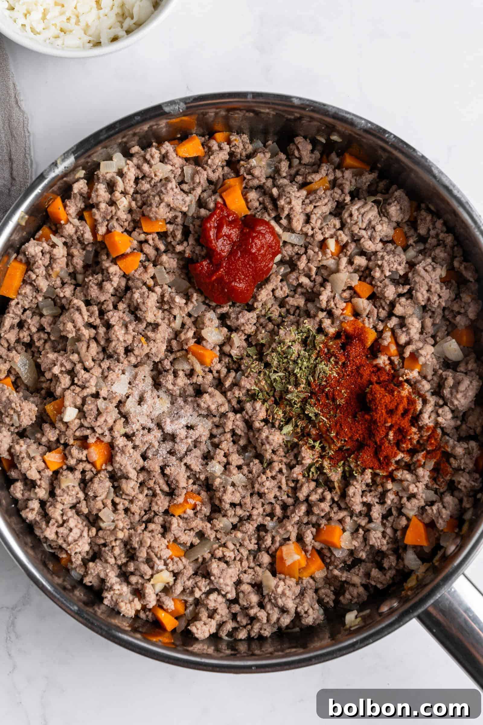 Crepe Lasagna Bake 8 A pan containing browned ground beef mixed with chopped onions and carrots. There's a dollop of tomato paste and sprinkled spices, including paprika and dried herbs, on top. The scene is set on a marble countertop.