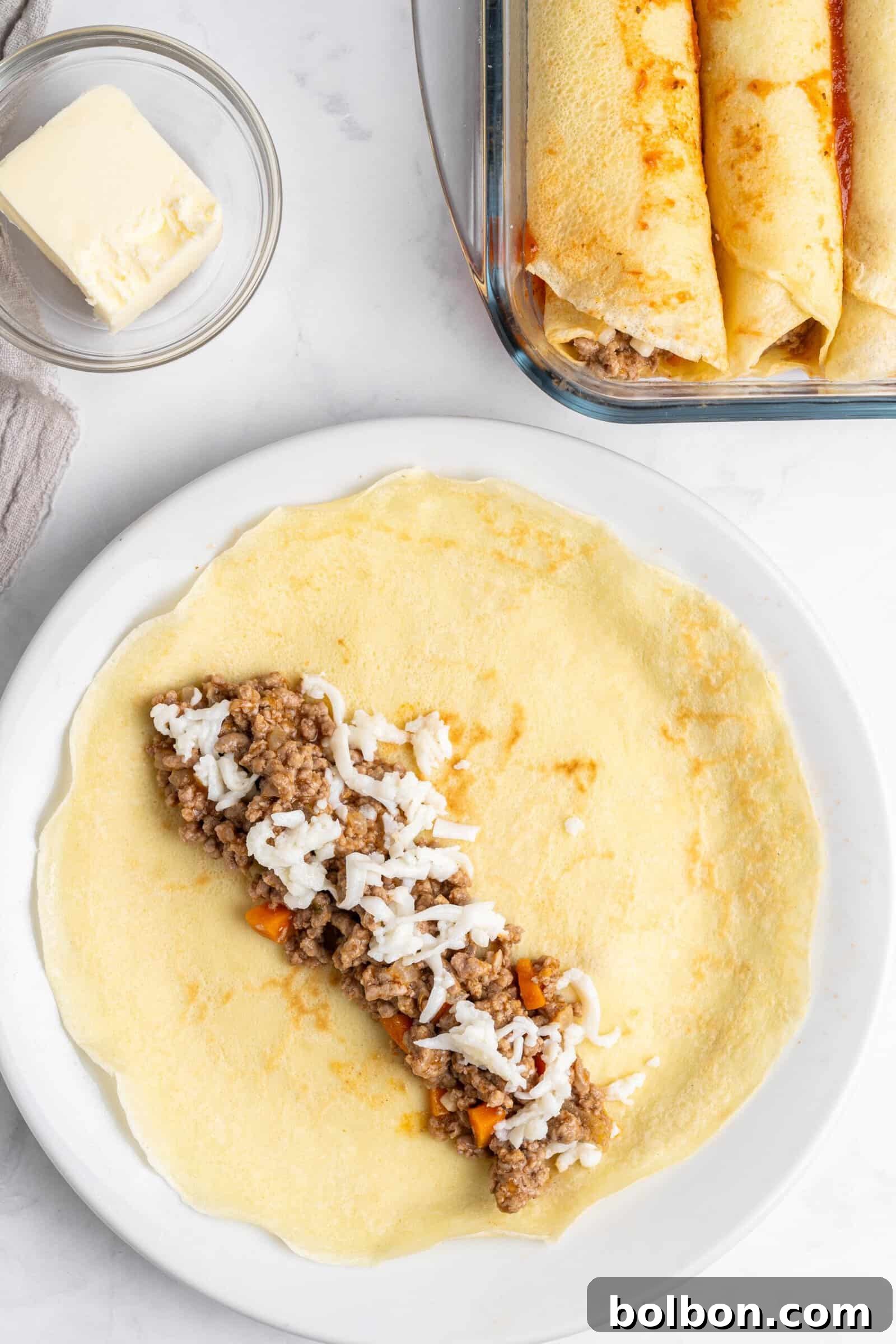 Crepe Lasagna Bake 9 A flat pancake on a white plate is filled with seasoned ground meat and shredded cheese, ready to be rolled. Nearby, a glass dish holds two rolled pancakes. A small bowl of butter sits beside a folded napkin on a marble surface.