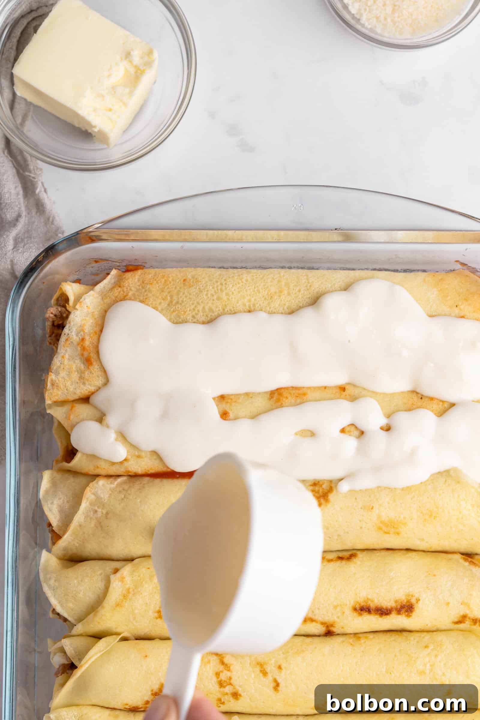Crepe Lasagna Bake 10 A baking dish filled with rolled crepes is being topped with a creamy sauce. A hand is pouring sauce from a measuring cup. Nearby, there is a dish of butter and a bowl of grated cheese.