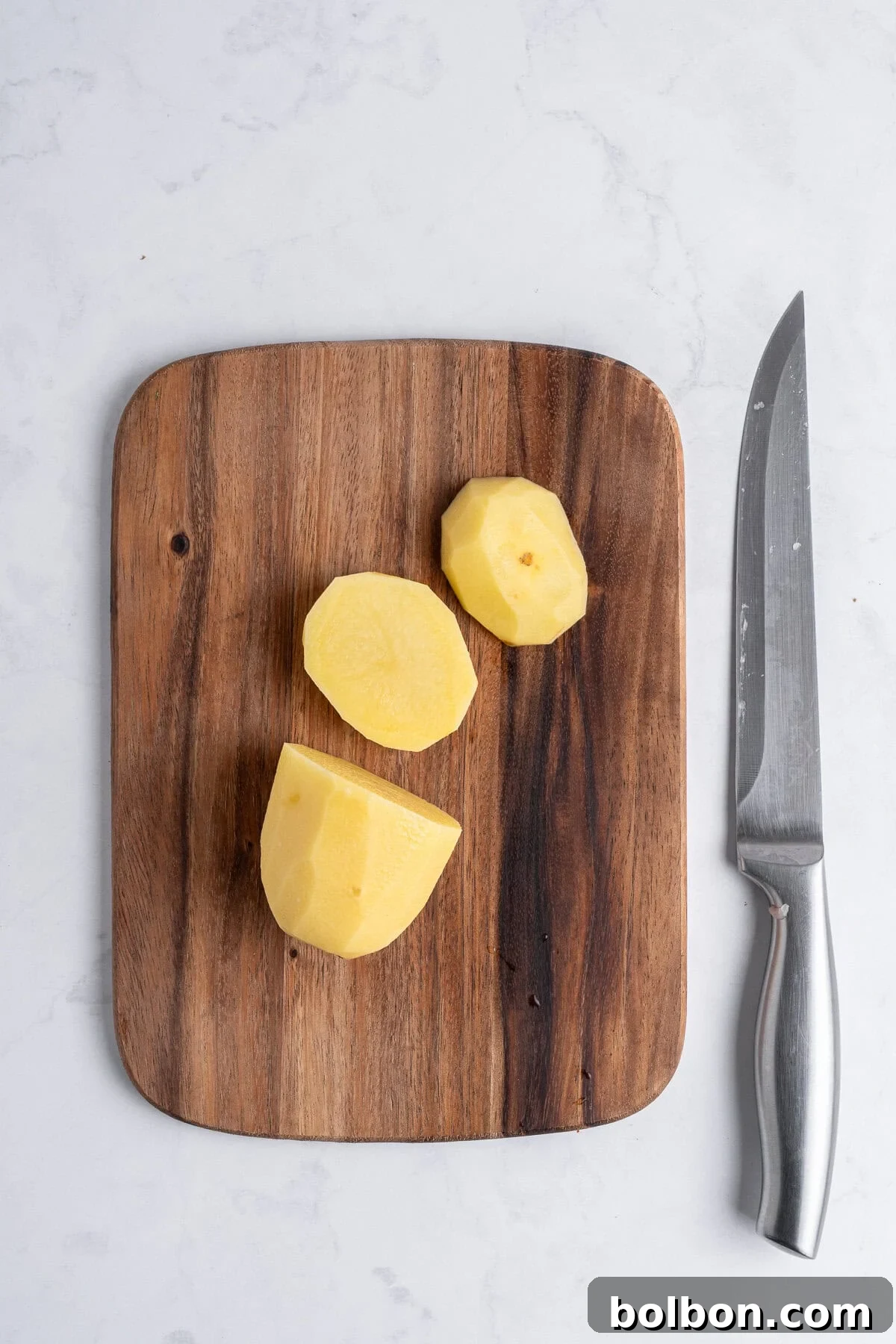A cutting board with a peeled, cut yellow potato on it and a large chef's knife laying beside it. 