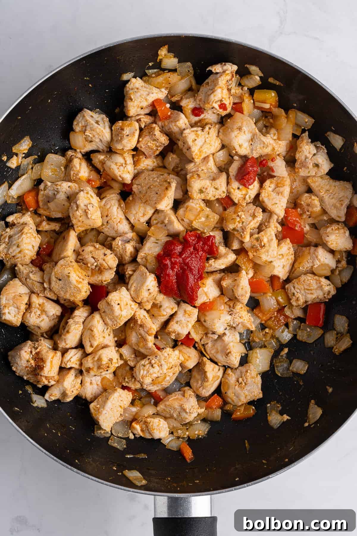 A saute pan with golden pieces of cooked, cubed chicken, as well as various spices adn tomato paste on top. 