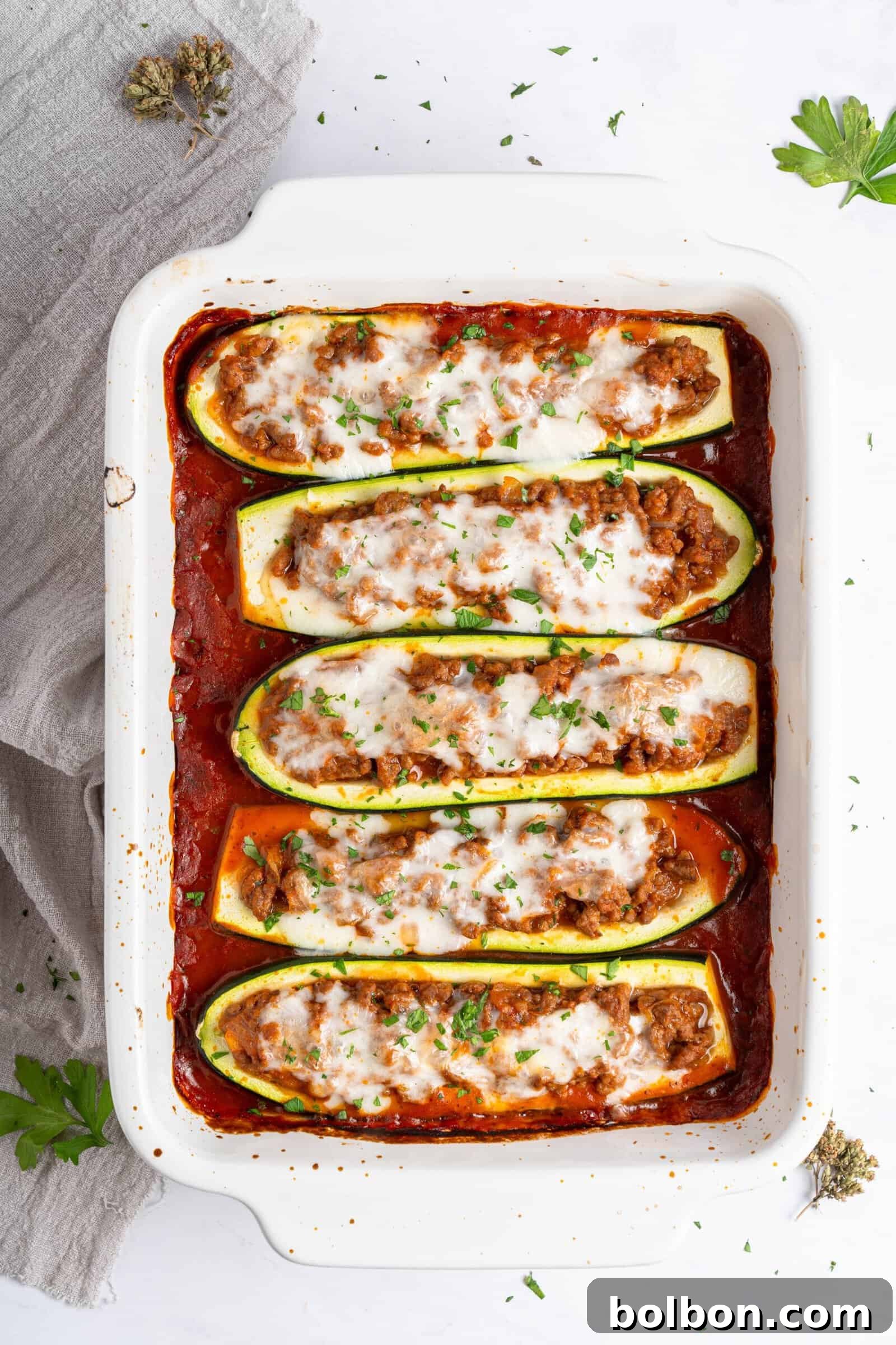 Loaded Zucchini Boats 2 A white baking dish with five zucchini boats filled with a meat mixture and topped with melted cheese, set in a bed of tomato sauce. The dish is garnished with chopped herbs and placed on a white surface with sprigs of herbs nearby, highlighting a perfect low-carb, easy weeknight meal.