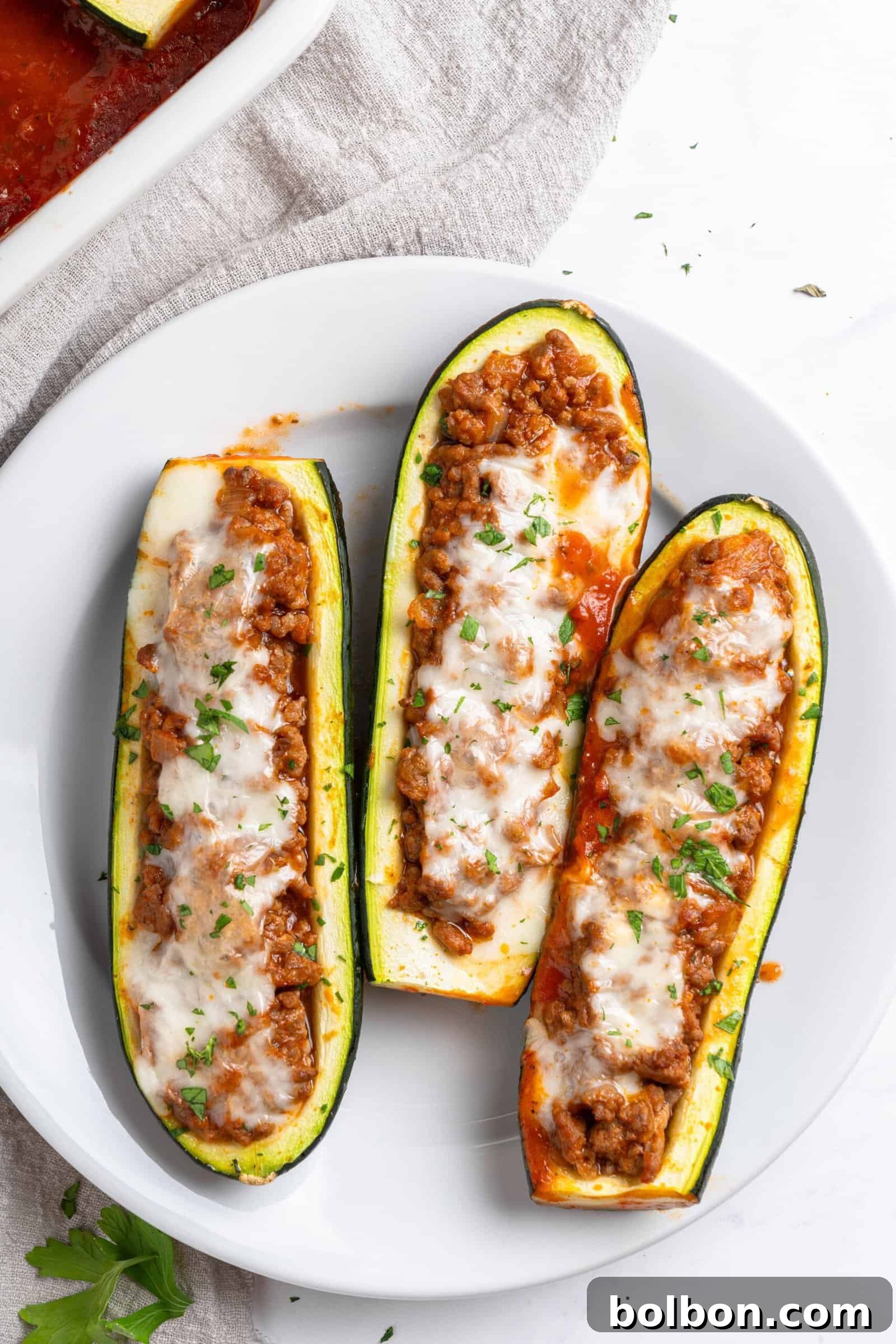 Loaded Zucchini Boats 12 Three zucchini boats filled with a seasoned meat mixture, topped with melted cheese and garnished with parsley, served on a white plate. The dish is placed on a light cloth, with part of a white baking dish visible in the background, showcasing a delightful family dinner.