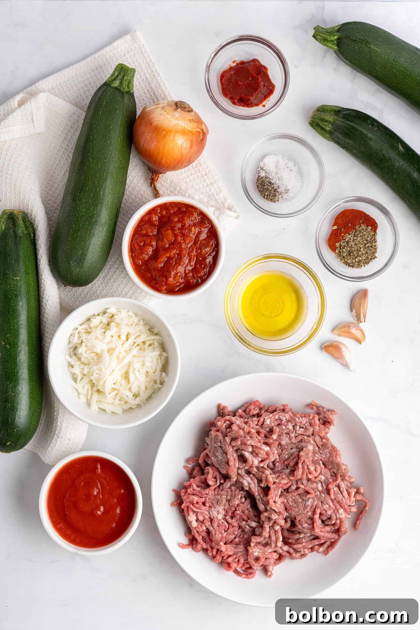 Loaded Zucchini Boats 3 Ingredients for a zucchini recipe arranged on a white surface: three zucchinis, ground meat, olive oil, tomato sauces, cheese, onion, garlic, red pepper flakes, oregano, salt, and pepper, showcasing fresh components for a healthy meal.