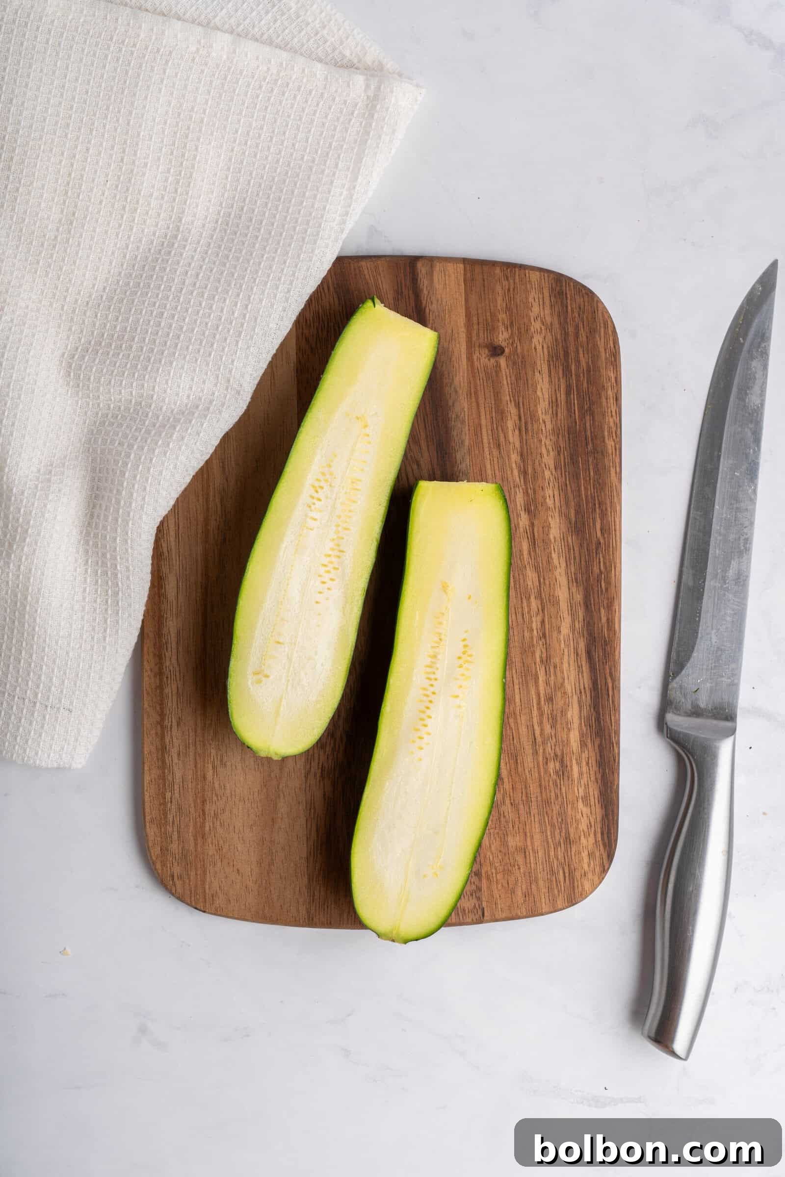 Loaded Zucchini Boats 4 Two zucchini halves are placed on a wooden cutting board, prepped for stuffing. A knife with a metallic handle lies to the right of the board, and a white textured cloth is partially visible to the left on a marble surface, illustrating the initial step of the recipe.