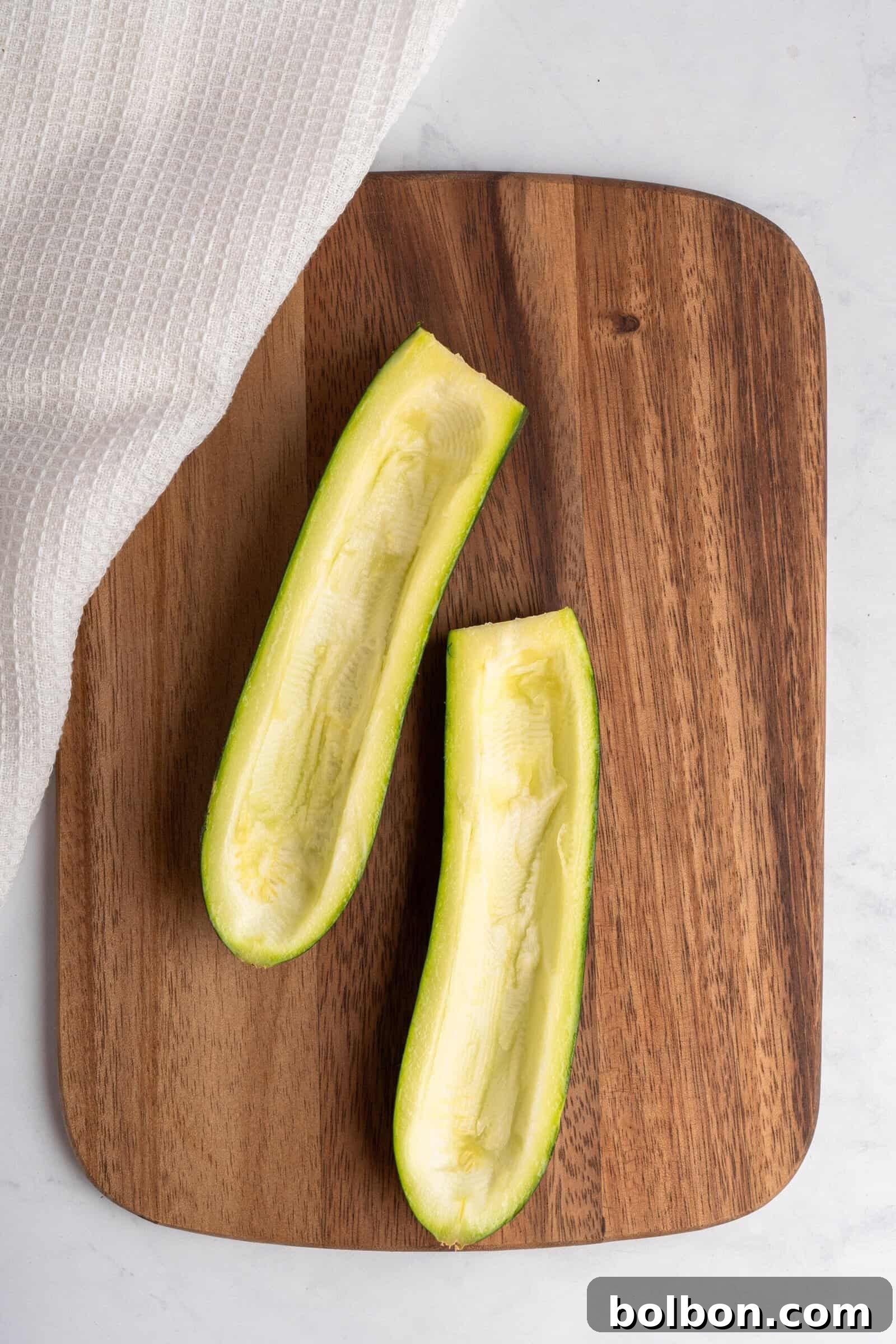 Loaded Zucchini Boats 5 Two zucchini halves with the seeds scooped out, resting on a wooden cutting board, ready to be filled. A white cloth partially covers the corner of the board, demonstrating proper preparation for zucchini boats.