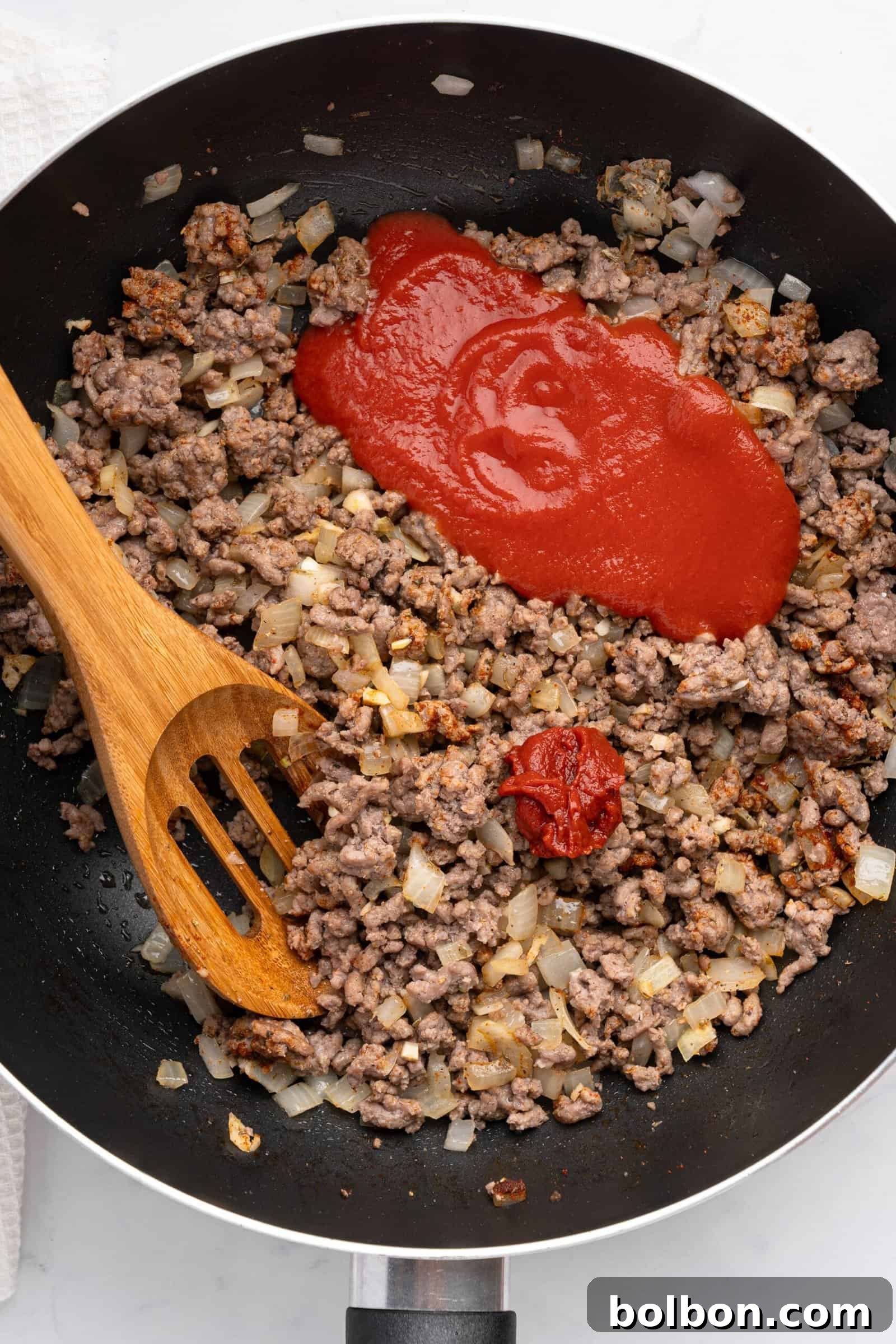 Loaded Zucchini Boats 7 A skillet contains browned ground beef and diced onions, topped with a splash of tomato sauce and a dollop of tomato paste. A wooden spatula rests in the pan, ready to stir the ingredients, highlighting the development of a savory meat sauce.
