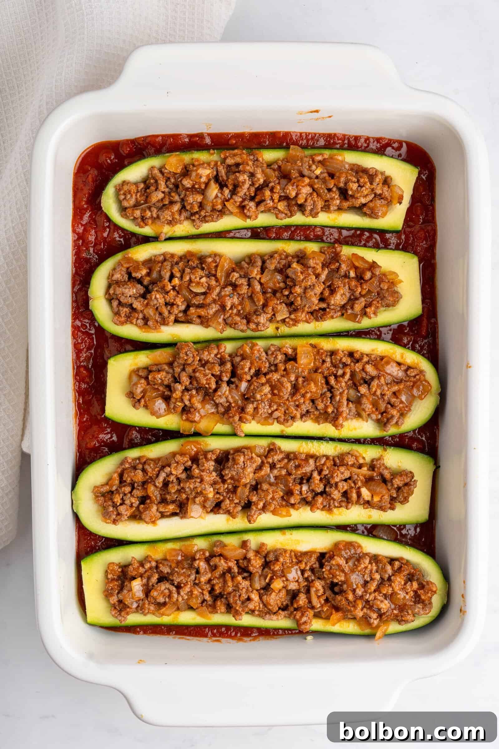 Loaded Zucchini Boats 9 Six zucchini boats filled with seasoned ground meat and vegetables are lined up in a white baking dish. The dish is placed on a light-colored surface. The zucchini boats are cooked in a tomato-based sauce, ready for the final step.