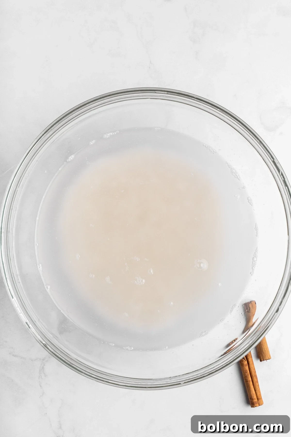 A clear glass bowl contains a light milky liquid on a white marble surface. Two cinnamon sticks are placed beside the bowl on the right.