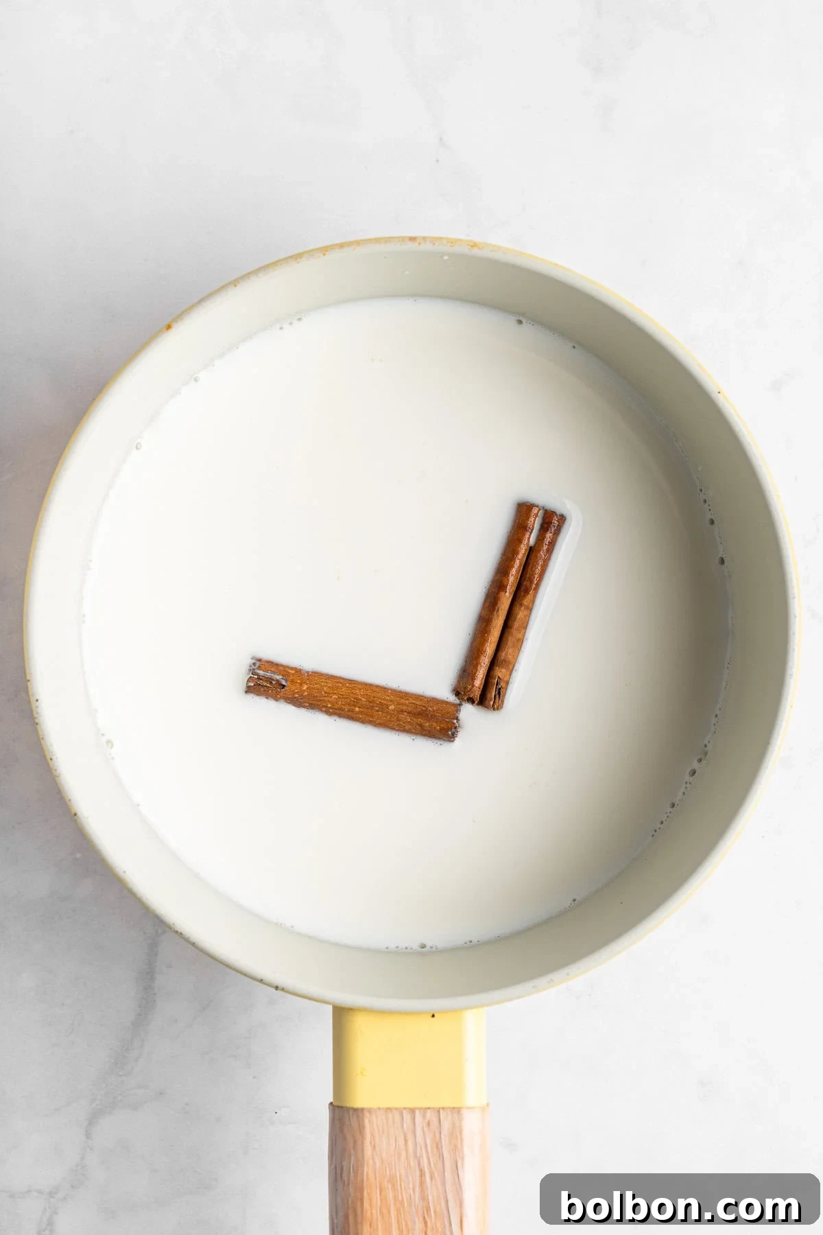 A saucepan filled with milk has two cinnamon sticks arranged to resemble a clock at 3 o'clock. The saucepan has a yellow exterior and a wooden handle, and rests on a light marble surface.