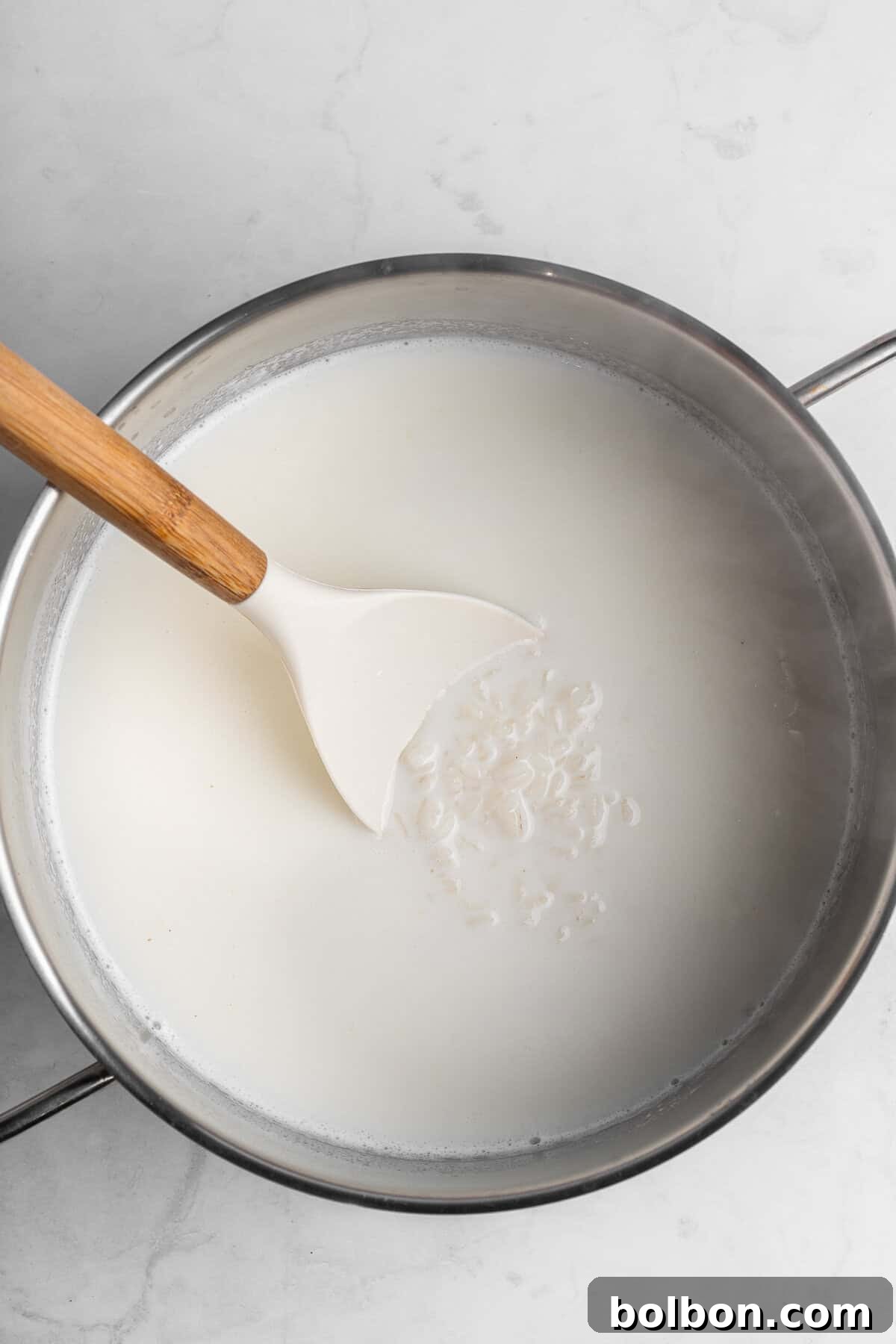 A pot filled with milk on a marble counter, with a white spatula stirring rice grains in the liquid.