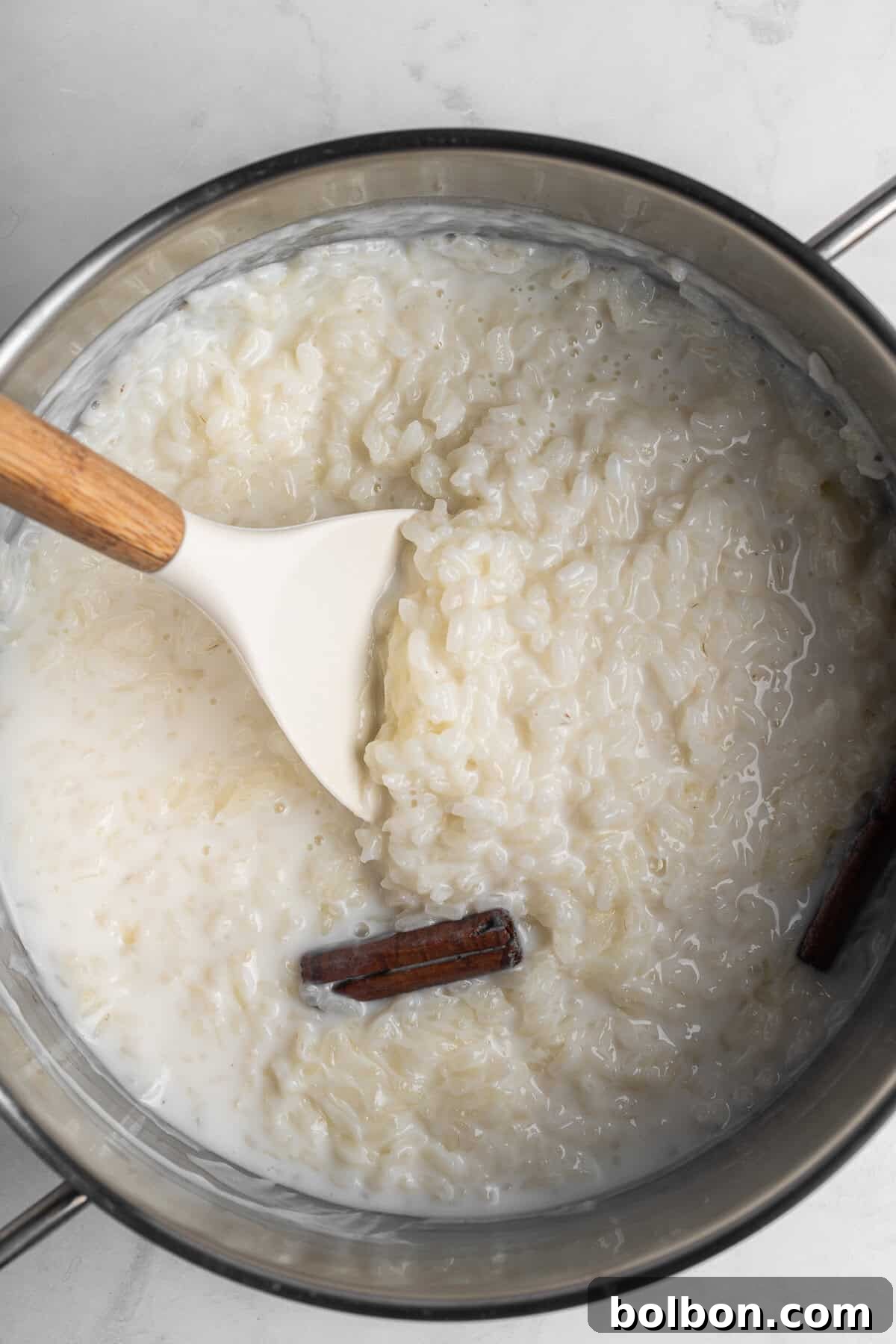 A pot of the creamy dessert. A cinnamon stick is partially submerged, and a white spatula with a wooden handle is resting in the pot. The background is a light marble surface.