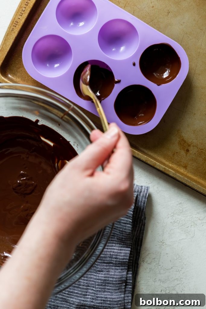 Crafting Irresistible Hot Chocolate Bombs 7 A spoon meticulously spreading melted chocolate inside a silicone sphere mold, ensuring an even and complete coating for a perfect hot chocolate bomb shell.