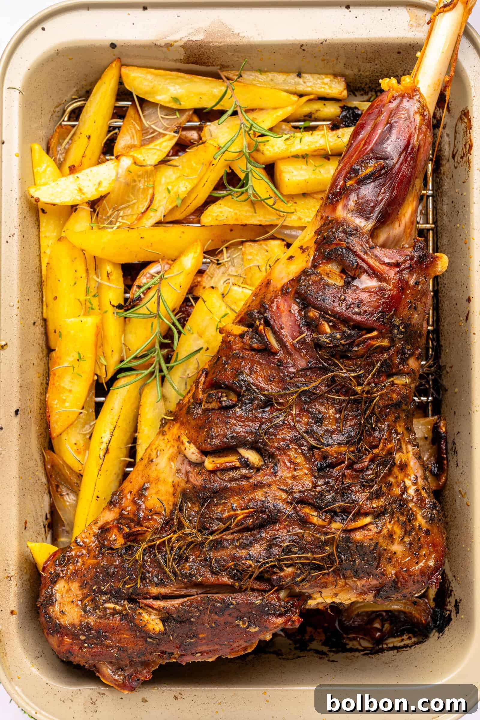 The Ultimate Roasted Lamb Leg 11 A roasted leg of lamb seasoned with herbs sits in a baking pan next to golden roasted potato wedges, garnished with sprigs of rosemary.